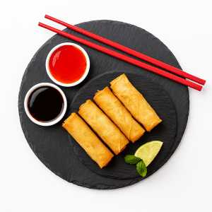 Plato redondo de pizarra natural de 25 cm con rollitos de primavera, dos cuencos de salsas y palillos chinos rojos. Presentación gourmet y elegante.